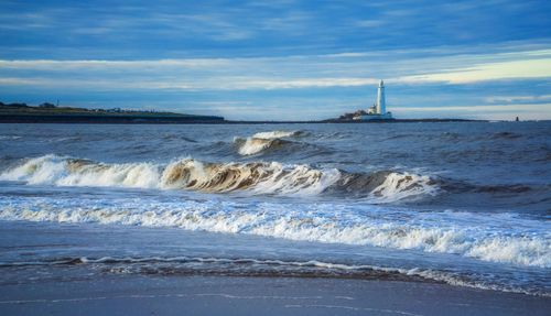 Free Majestic ocean waves crash against the seashore with a lighthouse standing tall in the distance. Stock Photo