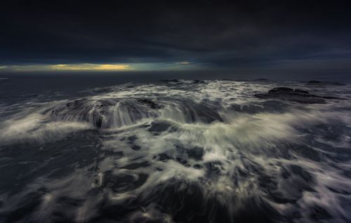 Free Powerful waves crash over dark rocks beneath a dramatic evening sky. Stock Photo