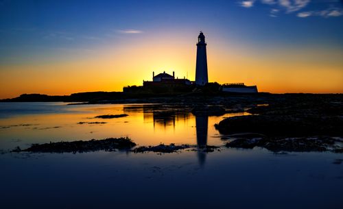 Free A stunning silhouette of a lighthouse against a vibrant sunset sky, reflecting beautifully in the calm sea. Stock Photo