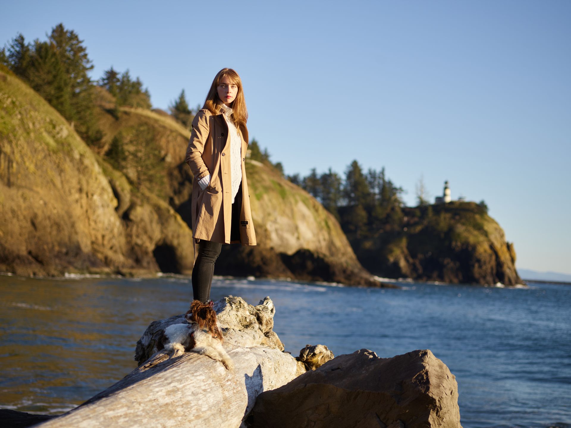 Caucasian woman standing on a log by the ocean with a distant lighthouse view.