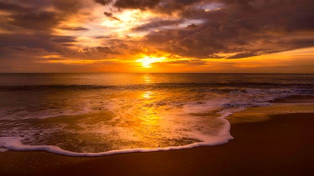 Golden sunset reflecting on the ocean waves and sandy beach with dramatic clouds.