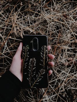 A hand holds a smartphone with botanical designs on dry grass, showcasing modern design.