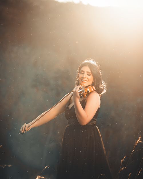 Free Smiling woman playing violin under sunlight, dressed in a black dress in outdoor setting. Stock Photo