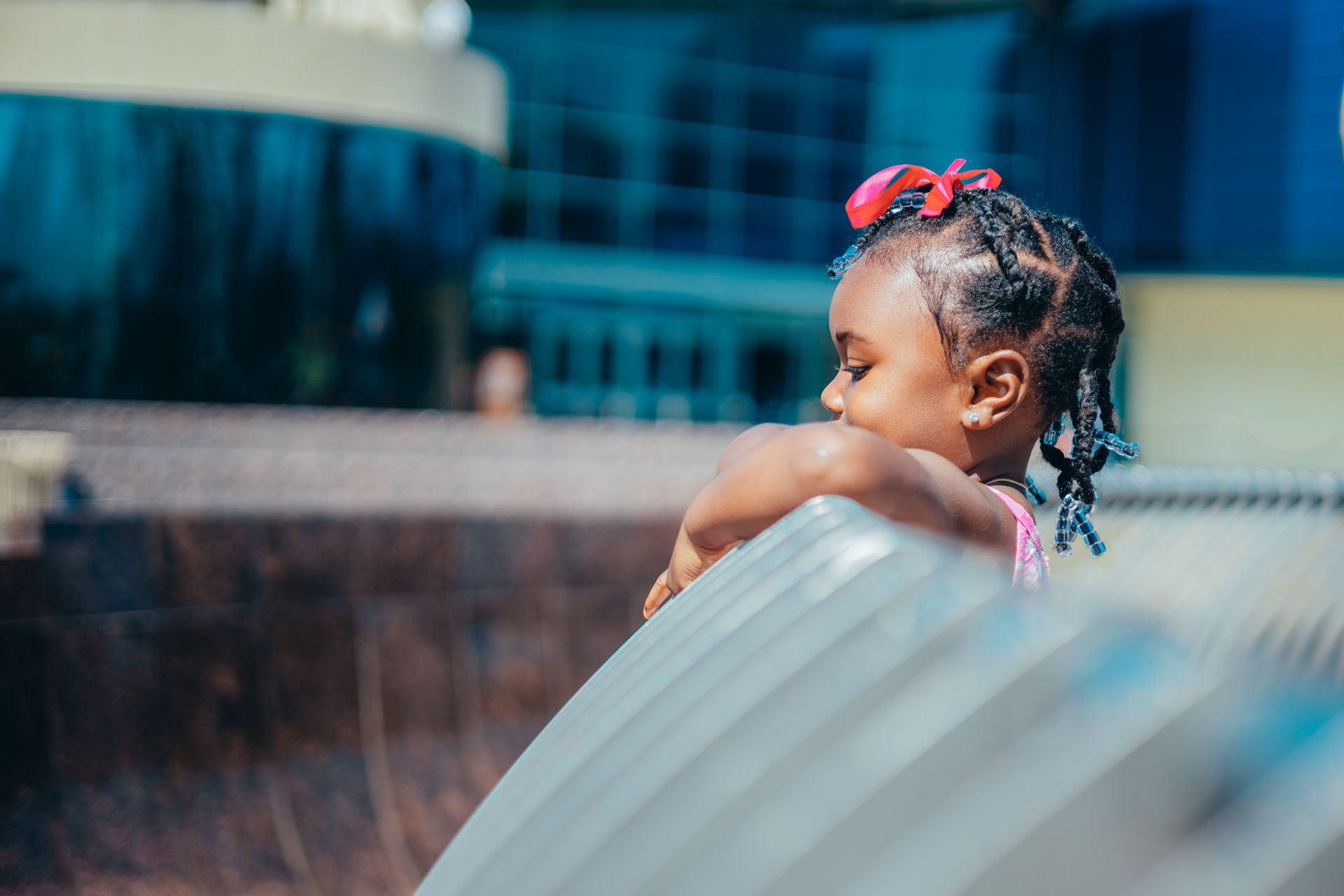 Adorable young girl with braided hair and a red ribbon, leaning on a railing outdoors.