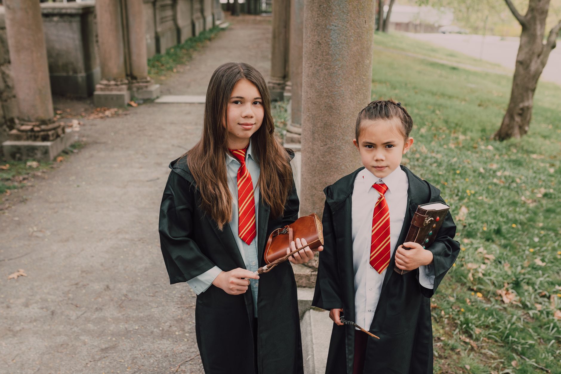 Two children dressed as wizards holding books and wands in a park, resembling a magical theme.