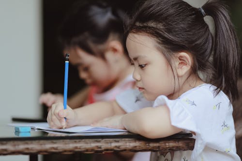 Free Adorable focused little Asian preschool girls sitting at table and doing tasks in copybooks in classroom Stock Photo
