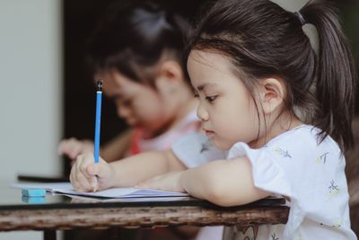 Adorable focused little Asian preschool girls sitting at table and doing tasks in copybooks in classroom