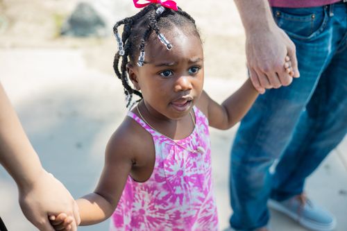 Free A cute child in a pink dress holding hands with adults, enjoying a sunny day outdoors. Stock Photo