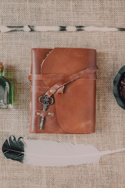 Flatlay of a leather notebook with vintage key, black feather, and green bottle on burlap fabric.