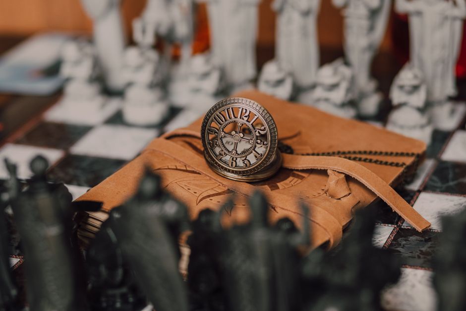 A steampunk clock rests on a leather-bound notebook with chess pieces in the background, evoking a magical atmosphere.