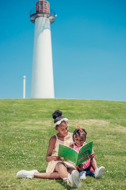 Free Two young girls enjoy reading together on a sunny day by a lighthouse, embodying happiness and sibling bonding. Stock Photo