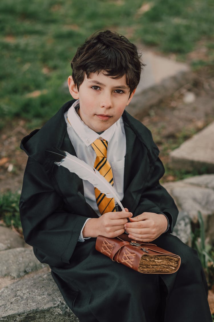 A Boy In Black Robe Holding A Spell Book And A Quill