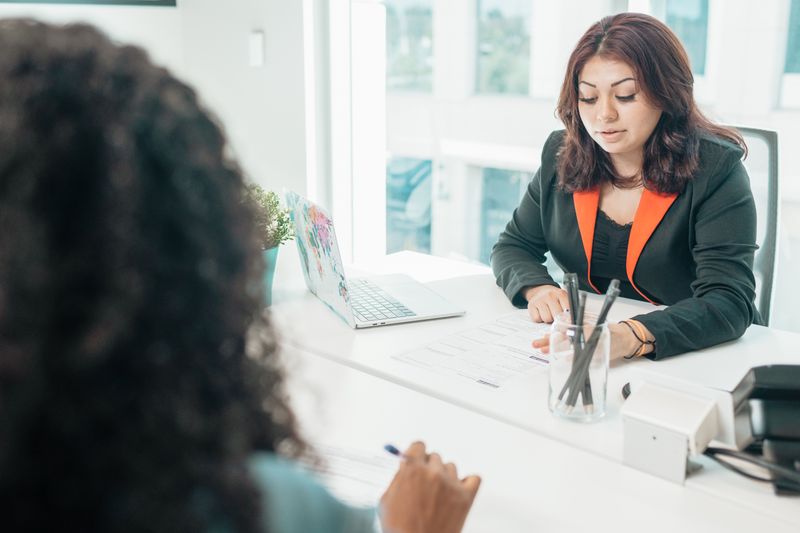 A person filling in a printed form at a counter with a blue pen, while a woman in a blazer looks on opposite.