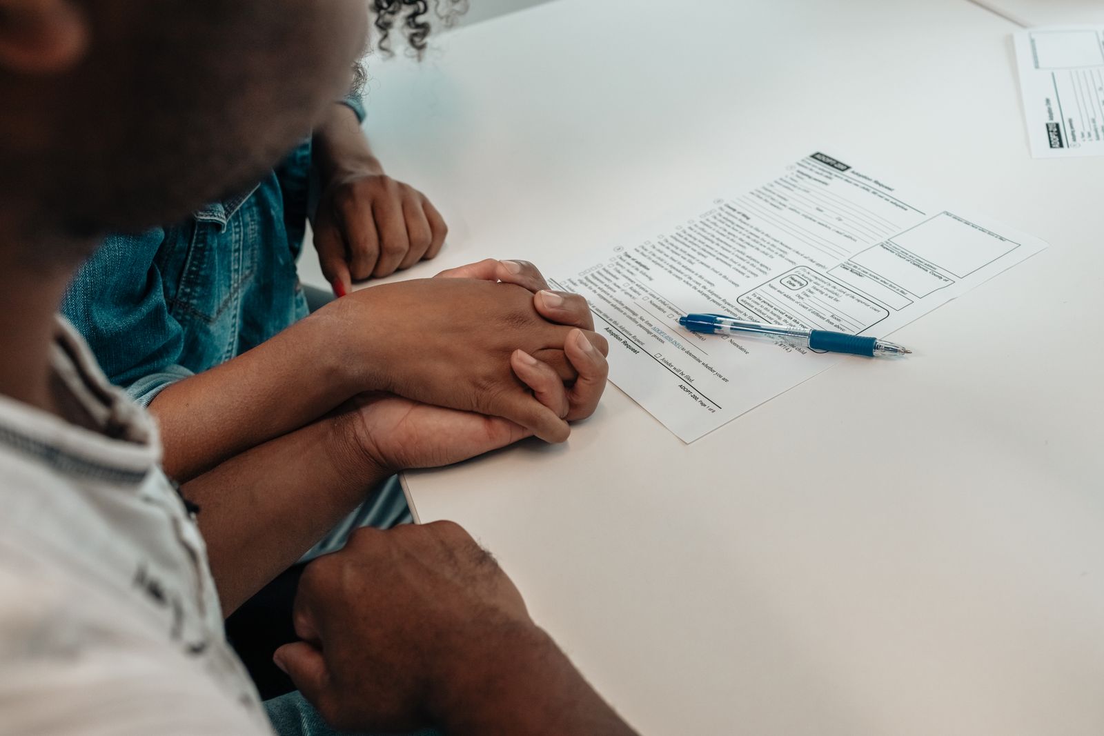 Two adults' hands hold a child's small hand over a printed form on a white table, a blue pen beside it.