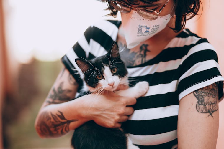 Woman wearing face mask holding a cute black and white kitten indoors with tattoos visible.