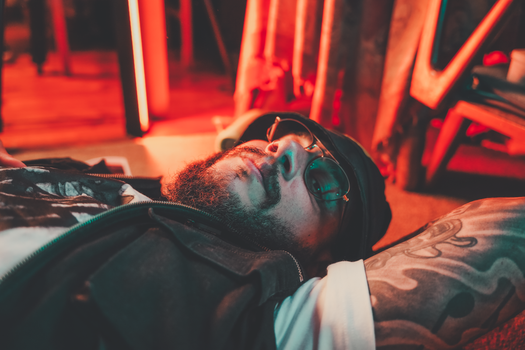 Close-up view of a tattooed man lying indoors under red lights, wearing a hat and glasses.