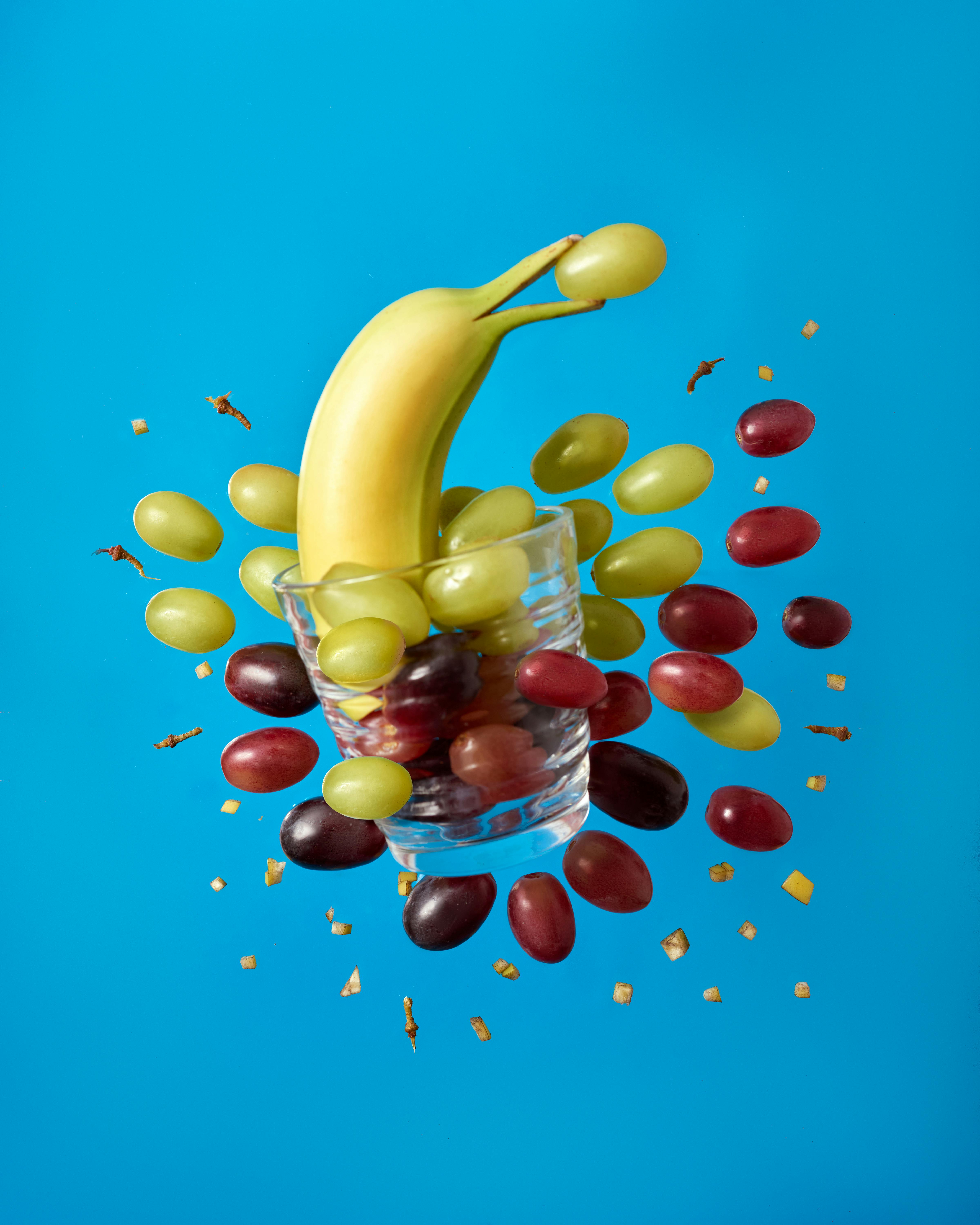A dynamic visual of a banana and grapes bursting from a glass on a blue background.