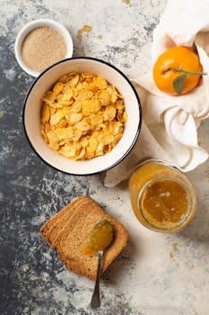 Top view of breakfast with cereal, toast, jam, and a mandarin on a rustic table.