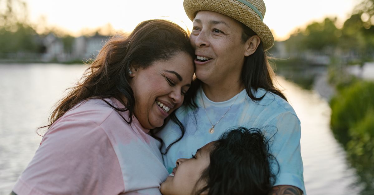 Happy same-sex family sharing an emotional hug outdoors near a serene lake at sunset.