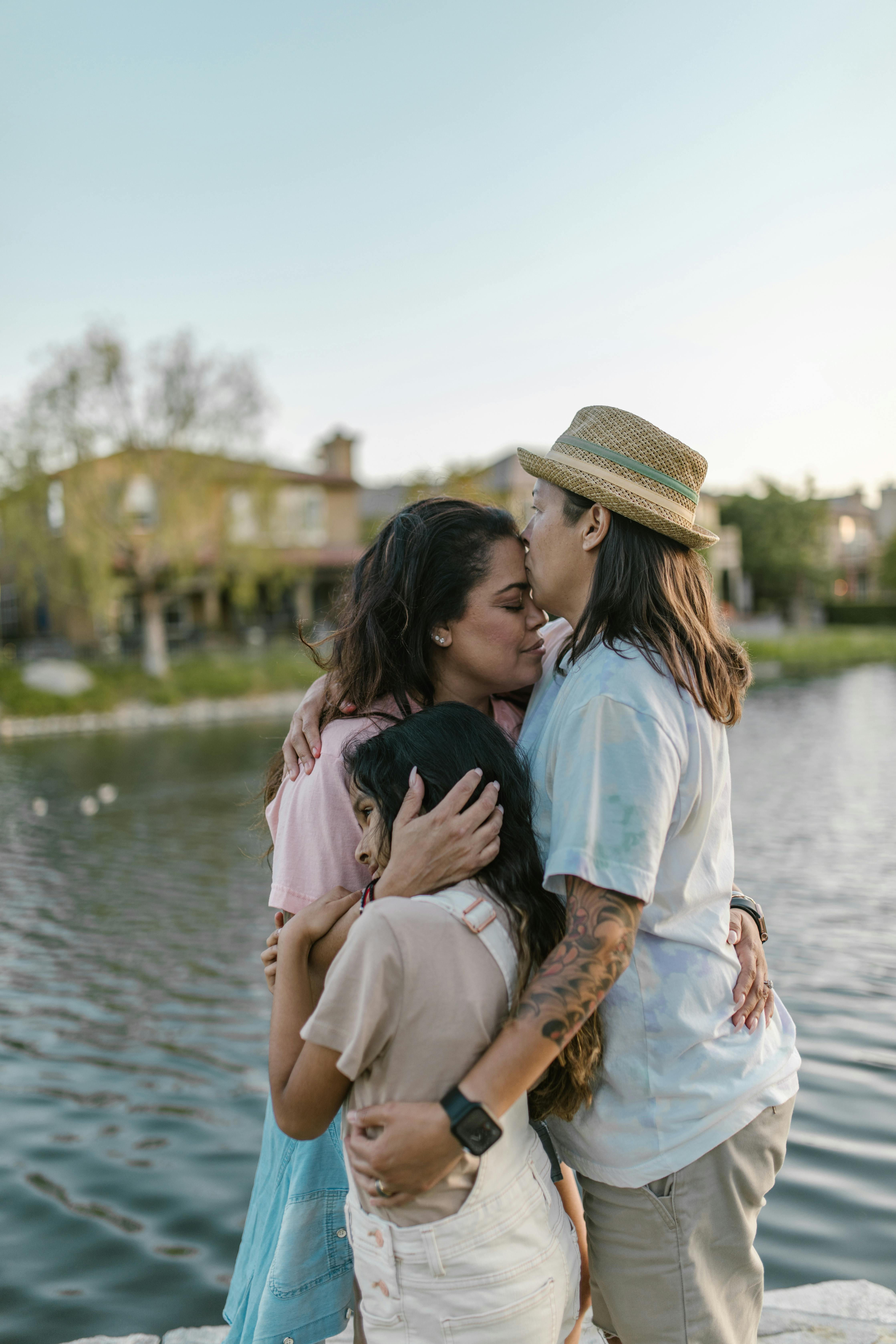 mothers kissing and embracing their daughter while standing by a body of water in summer