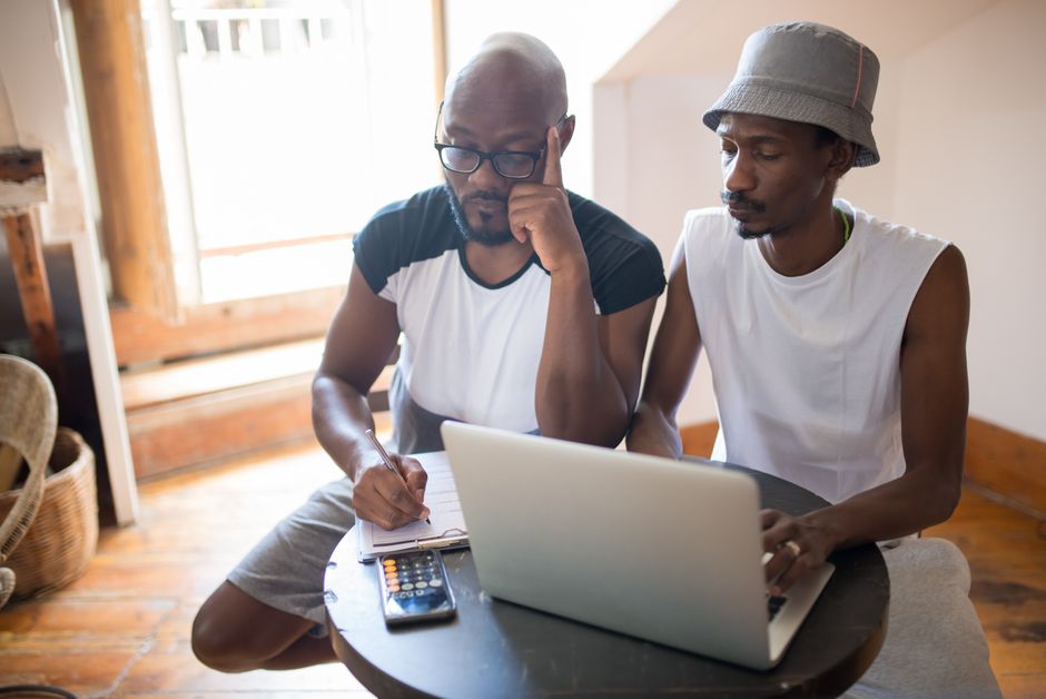 Two men collaborating indoors with a laptop and clipboard, showcasing teamwork and focus. Two men collaborating indoors with a laptop and clipboard, showcasing teamwork and focus.