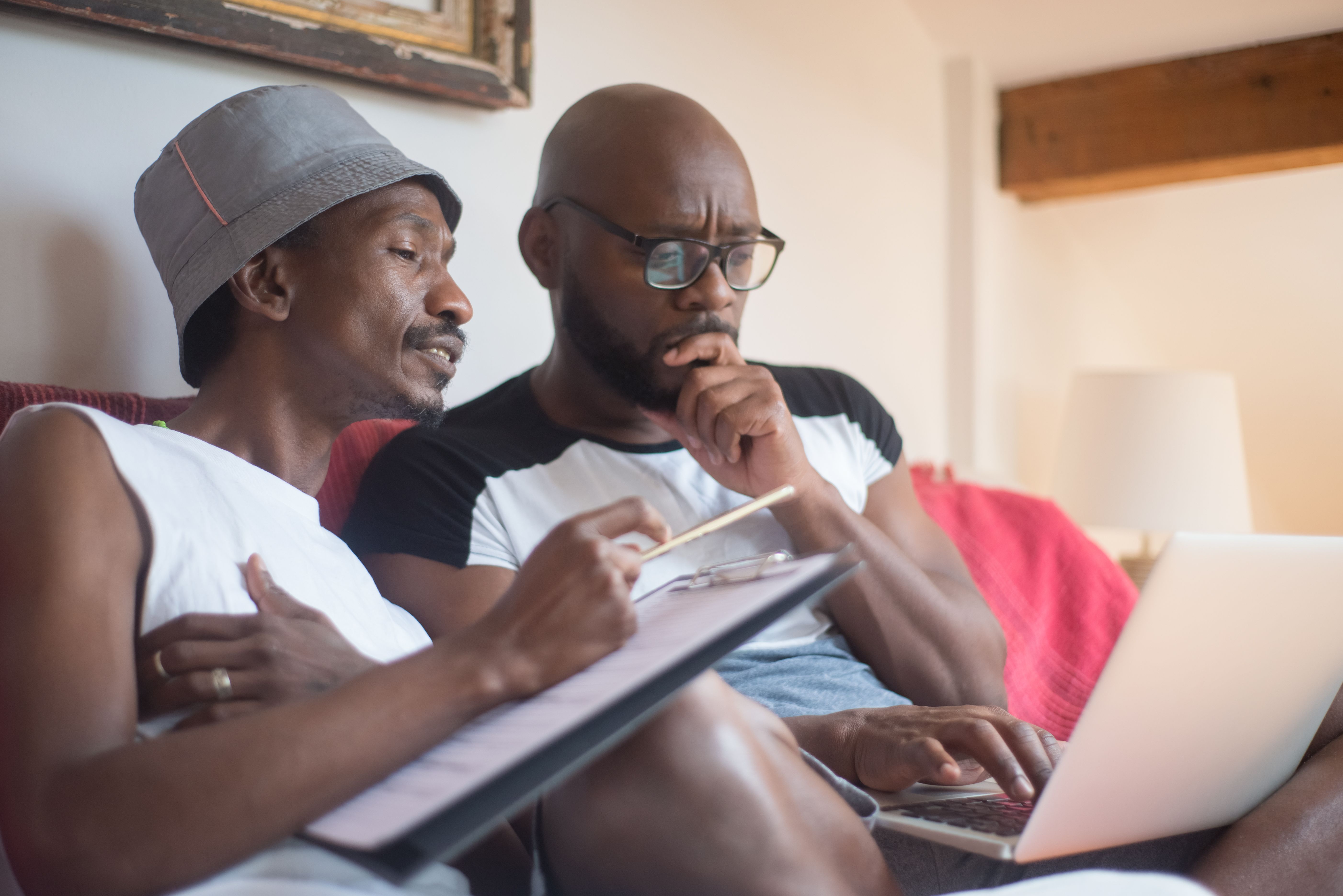 Two men discussing work on a sofa with a clipboard and laptop, thinking deeply.
