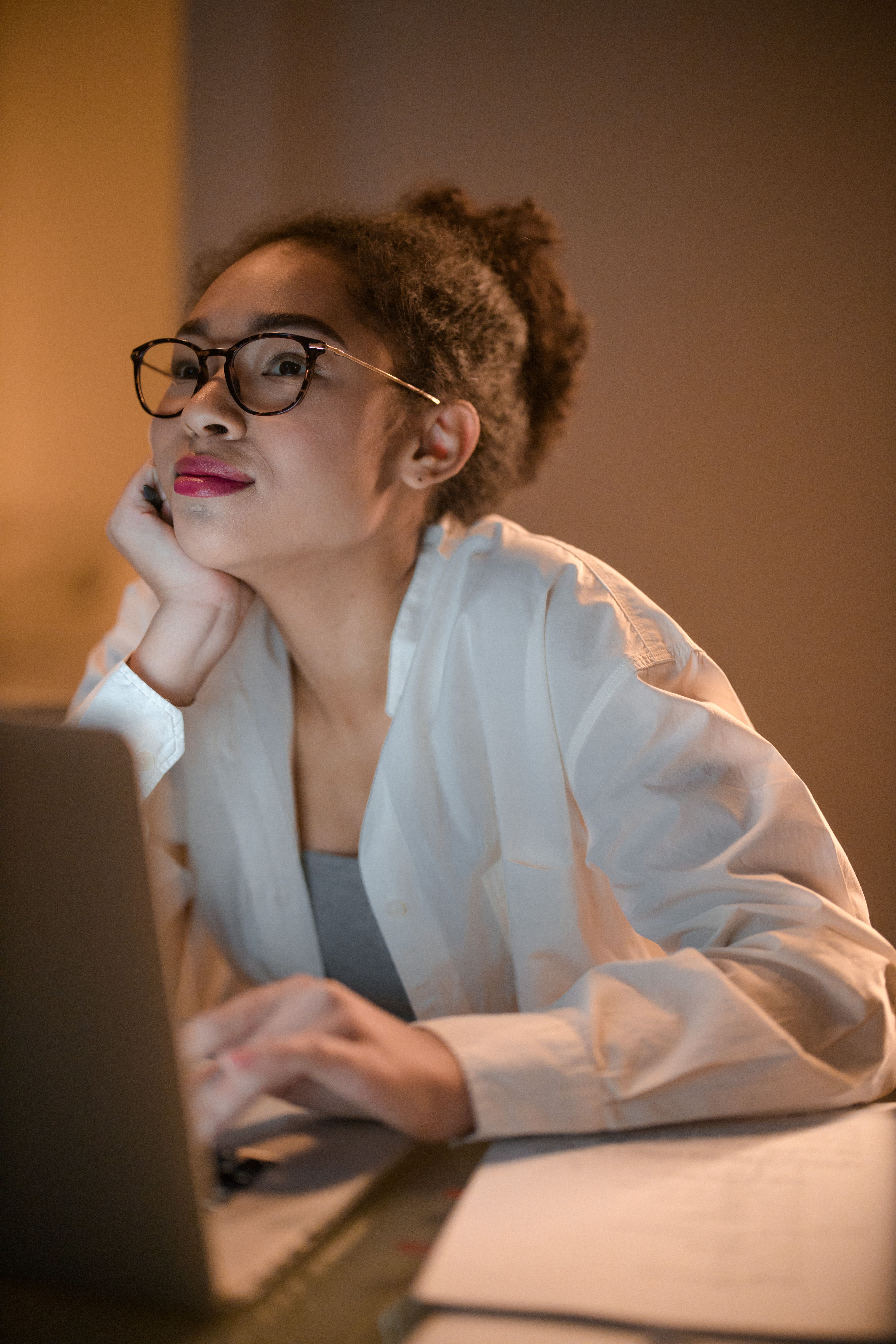 A young woman in glasses works on her laptop at home, contemplating her thoughts.