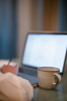 A ceramic coffee mug in focus with a blurred laptop and writing hand in the background.