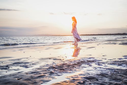 Free A serene scene of a woman walking along the beach at sunset, reflected in the water. Stock Photo