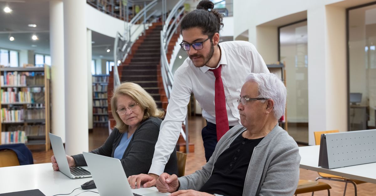 A young mentor guides senior adults using laptops in a modern library setting, fostering digital literacy.