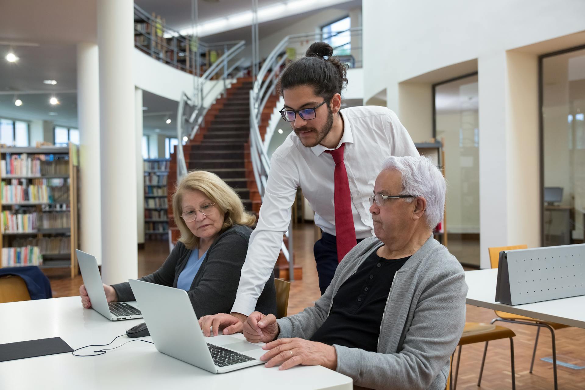 A young mentor guides senior adults using laptops in a modern library setting, fostering digital literacy.