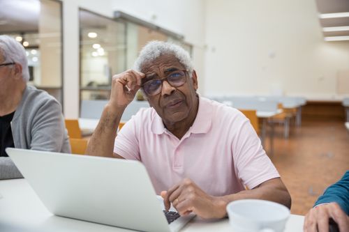 Free Elderly man in glasses sitting at a table, using a laptop in a bright, modern room. Stock Photo