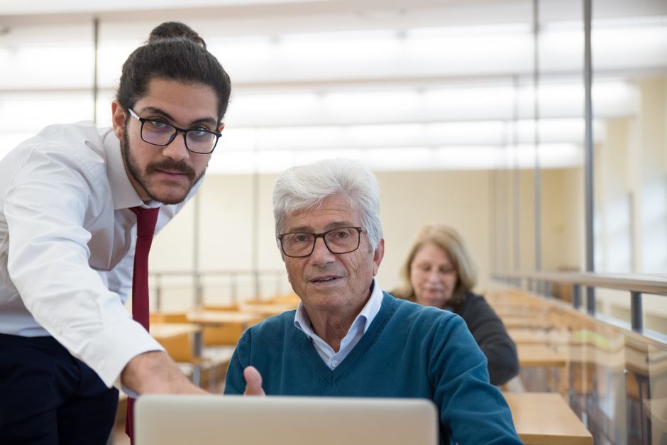 benefits of continuous learning for senior professionals - An elderly man learning computer skills with a teacher in a classroom setting, showcasing education through technology.