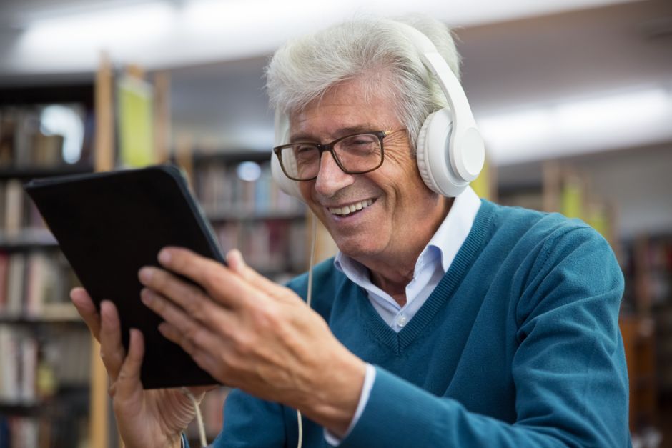 Elderly man happily using tablet with headphones in a library setting.