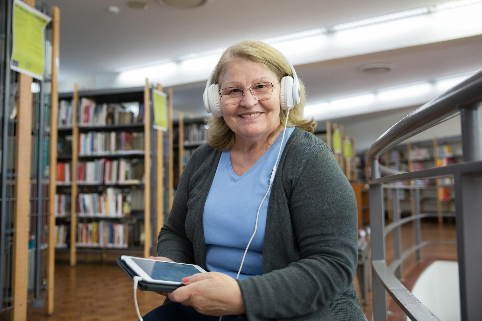 Senior woman smiling while listening to audiobooks on tablet in library setting.