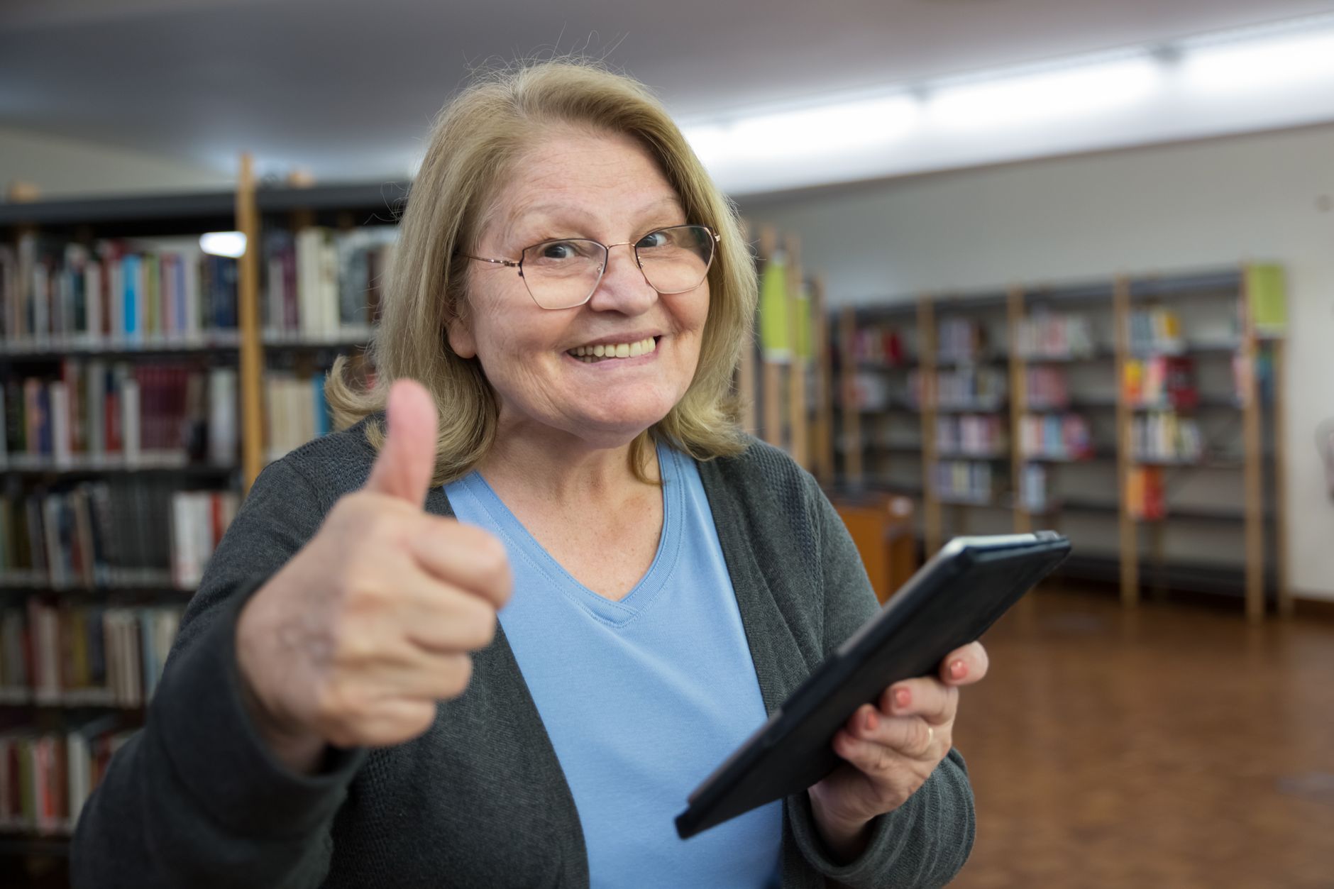 為何每個人都該具備數位素養?生活工作全面指南 Happy senior woman gives a thumbs up while using a tablet in a library, embracing technology.