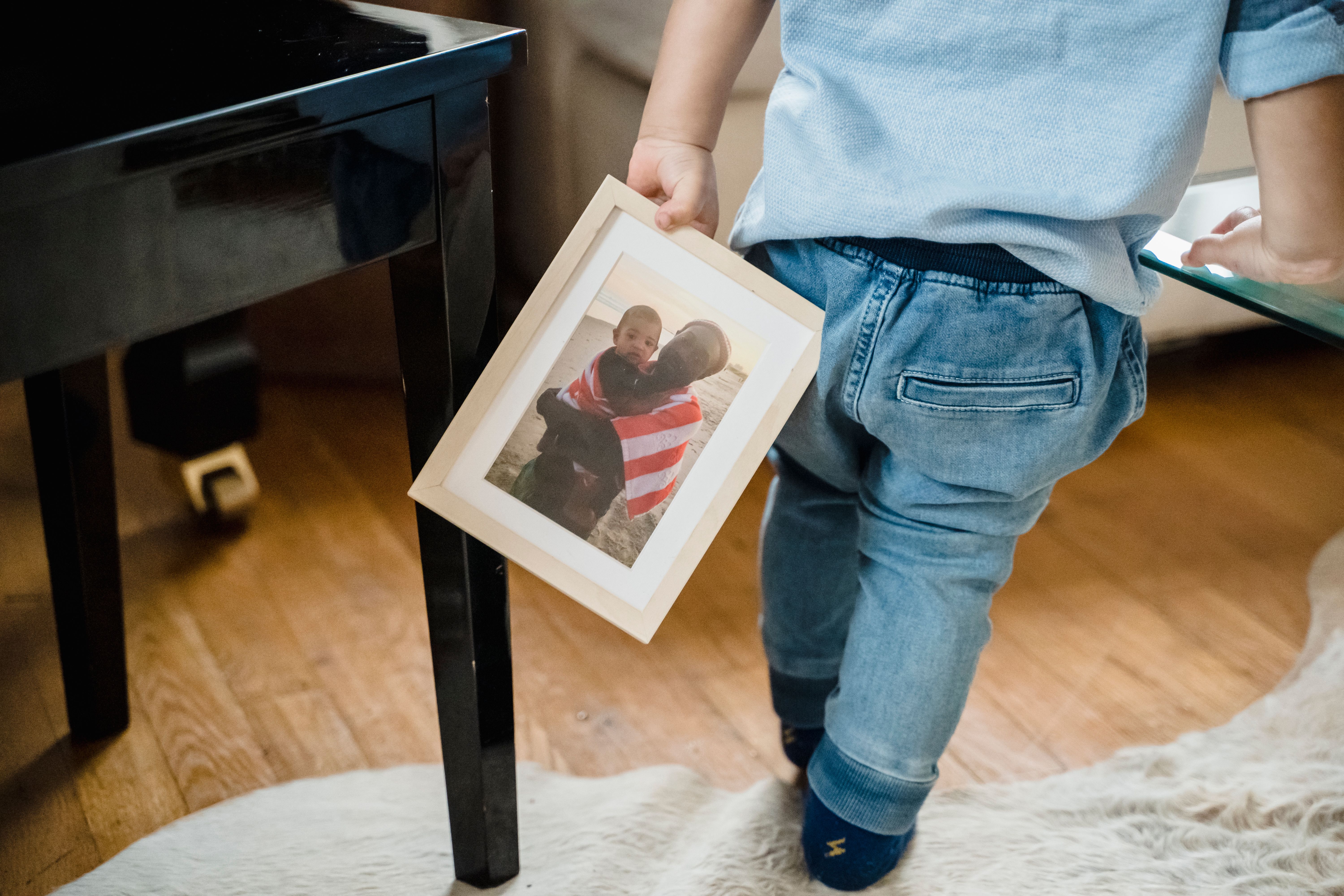 A toddler walking indoors, holding a framed family photograph, capturing a moment of togetherness.