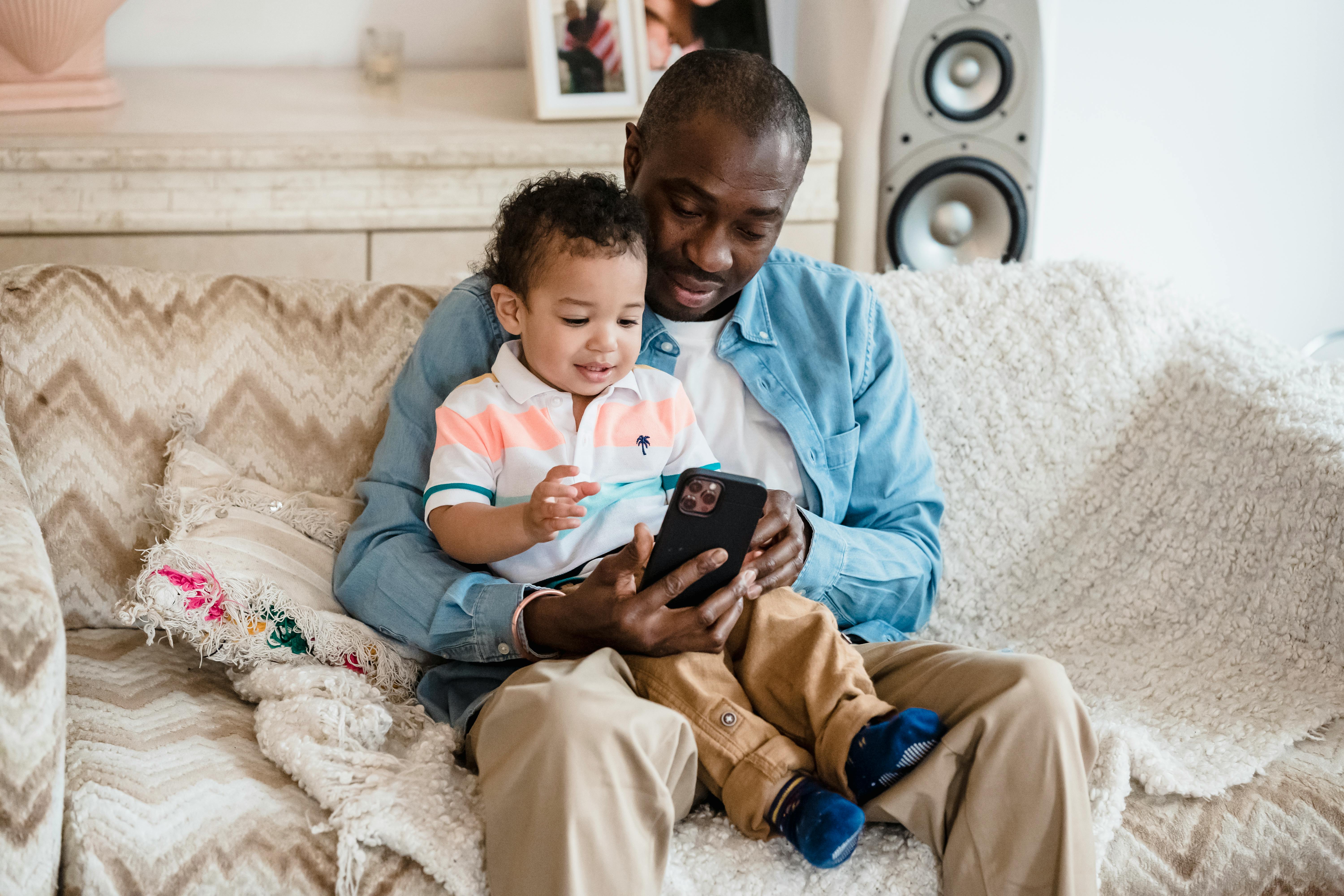 Free Man Sitting on a Sofa with his Son and Playing a Game on a Phone Stock Photo