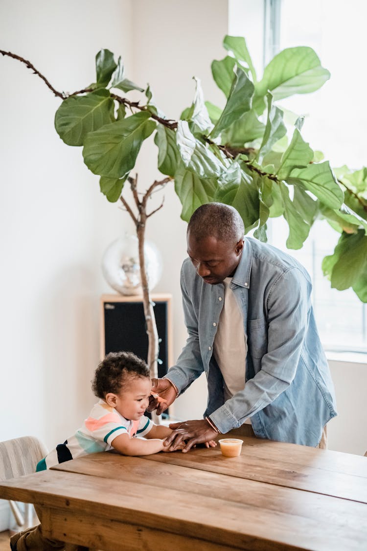 Father Sitting Toddler Down By Table For Snack