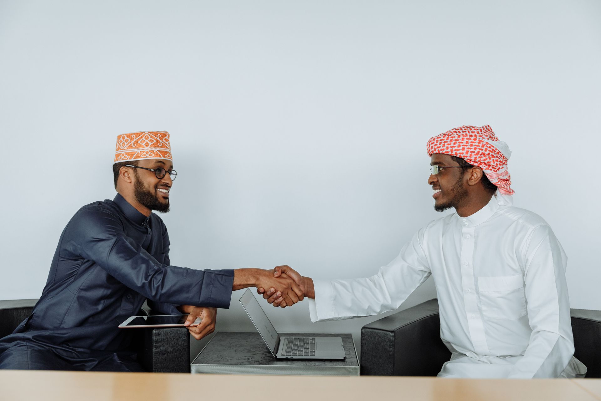 Two businessmen in traditional attire shaking hands in a modern office with laptops.