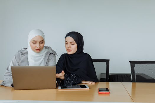 Two Muslim businesswomen in hijabs working together on a laptop in an office setting.
