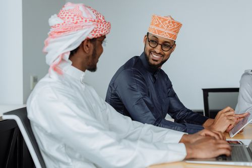 Free Men in traditional attire collaborating on laptops in an office, smiling and engaging. Stock Photo