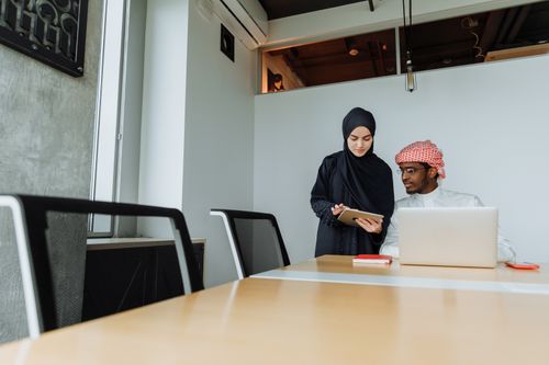 Free A man and woman in traditional clothing collaborate in a modern office setting, using tablets and laptops. Stock Photo