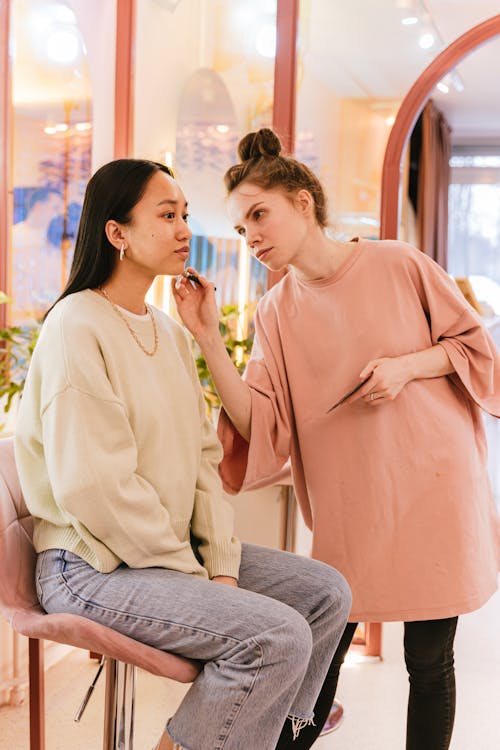 Woman in Peachy T-Shirt Doing Makeup on Woman Sitting on the Chair