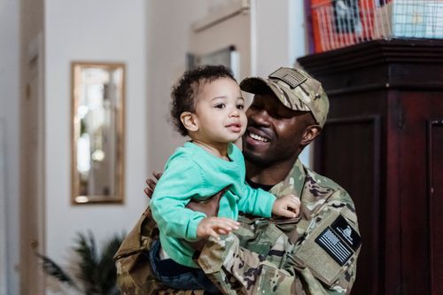 Free A cheerful father in military uniform holds his young child indoors, showcasing parenting and family bonds. Stock Photo