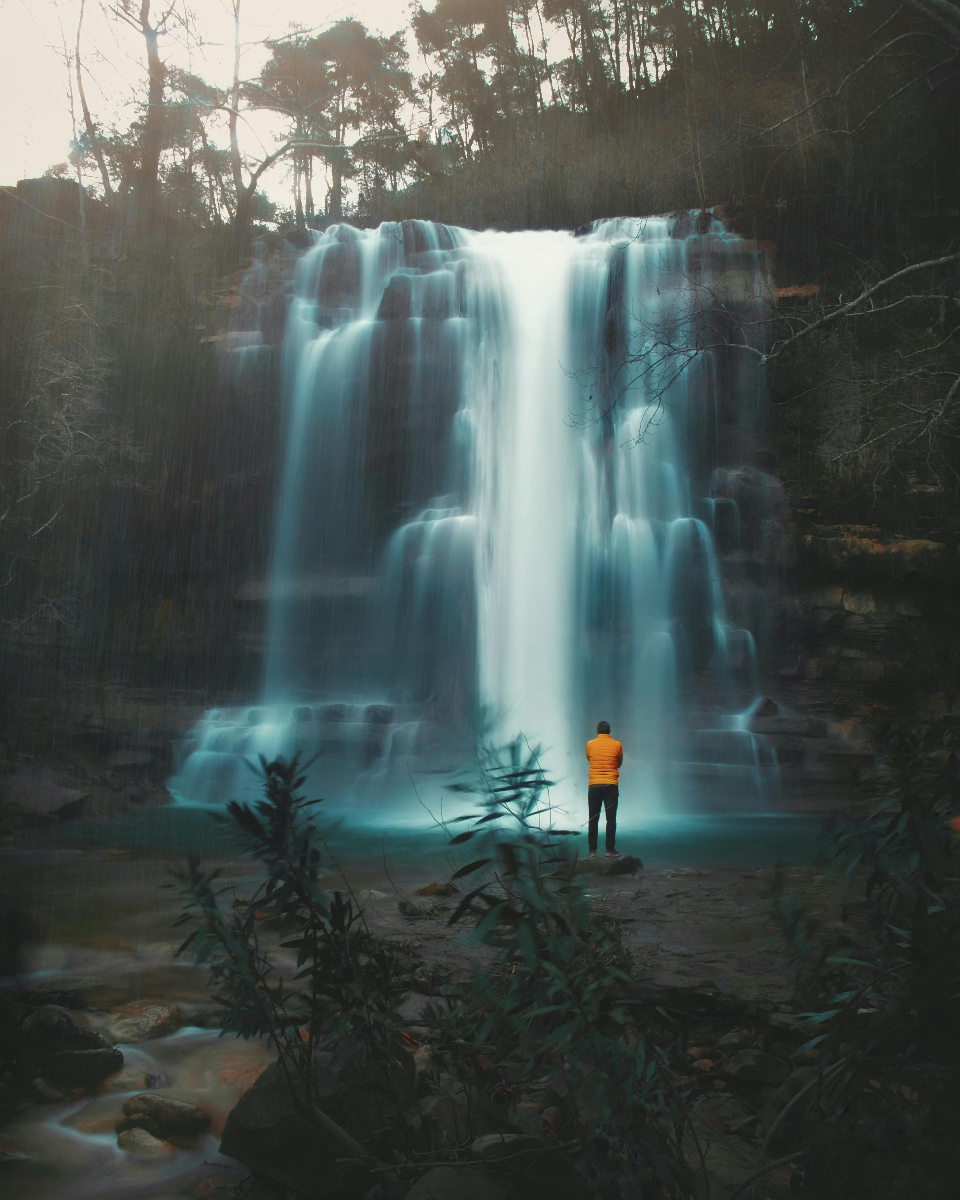 Back view of anonymous male traveler admiring picturesque waterfall cascade flowing down rocky terrain covered with green trees