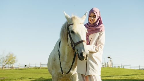 Free A young Muslim woman wearing a hijab, smiling and standing with a white horse in an outdoor setting. Stock Photo