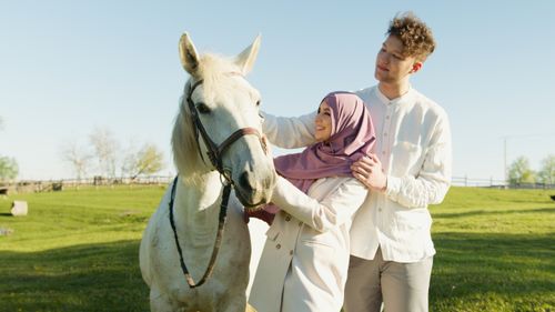 Free A smiling couple bonding with a white horse on a sunny day outdoors in a lush field. Stock Photo