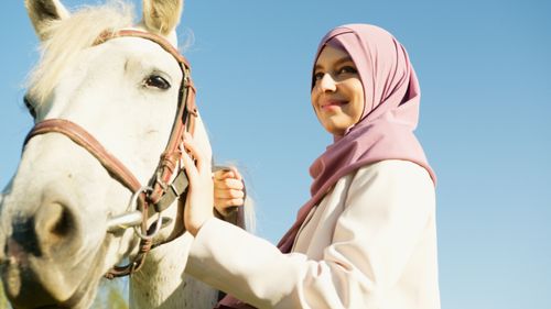 Free A Muslim woman in a hijab smiles while standing next to a horse on a sunny day. Stock Photo