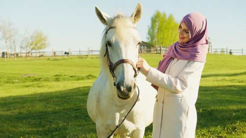 Free A young woman in a hijab smiles while petting a white horse in a sunny outdoor field. Stock Photo